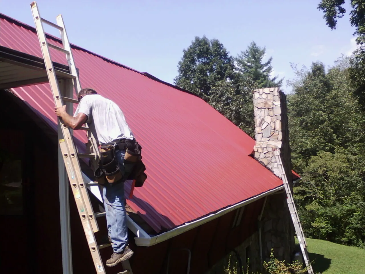 Roofer on a ladder beside a red metal roof and stone chimney — Soffit & Fascia Repair by Roofing Brothers in Old Fort